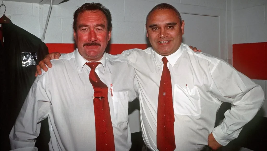 Two men in white shirts and red ties, standing together.